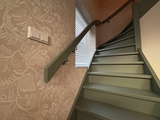 Behangen van trapgat en schilderen trap Groene trap met leuning, tegen een muur met behang en een raam aan de bovenkant.